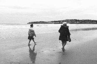 Two people walking barefoot along a quiet beach shoreline on a cloudy day, with a town visible in the distance