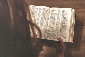Person reading an open Bible at a wooden table, viewed from overhead with softly lit pages