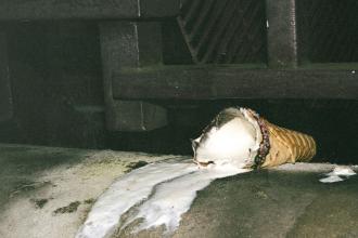 A dropped ice cream cone lies on the ground, with melted ice cream spreading across the surface near a dark metal structure