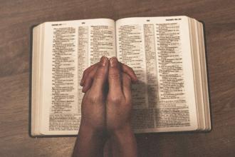 Hands folded in prayer over an open Bible on a wooden surface