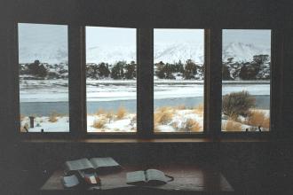 Open Bibles on a wooden table inside a cabin, overlooking a snowy landscape and frozen river through large windows