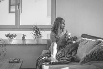 Person relaxing on a sofa with a blanket and holding a warm drink near a bright window with potted plants