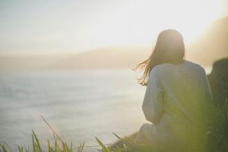Person sitting in the grass overlooking a calm body of water at sunrise, with sunlight creating a soft, peaceful glow