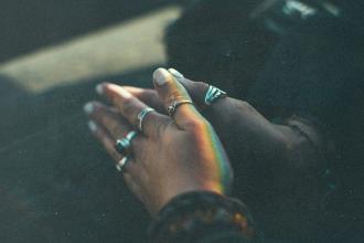 Hands clasped together in soft light, adorned with silver rings and touched by a faint rainbow reflection