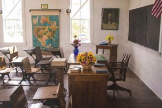Vintage-style classroom with wooden desks, a teacher’s desk decorated with flowers, a large map on the wall, and sunlight streaming through tall windows