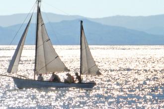 Sailboat carrying several people gliding across a sunlit lake with sparkling water and distant mountains in the background