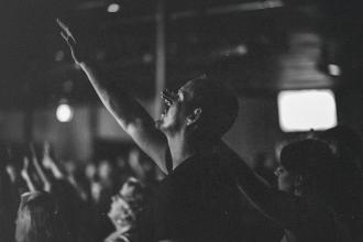 Person raising an arm in worship during a dimly lit church or concert gathering with others in the background