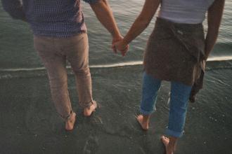Two people holding hands while walking barefoot along the shoreline where the water meets the sand