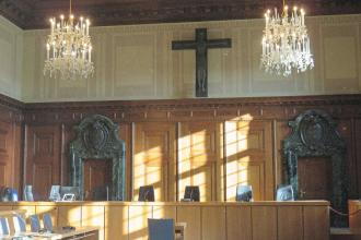 Elegant courtroom with wooden paneling, chandeliers, and a large cross mounted on the wall above the judges’ bench