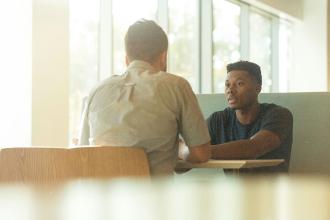 Two people sitting across from each other at a table in a bright room with large windows, engaged in conversation