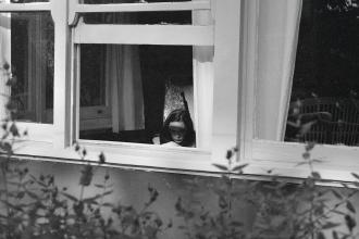 View through an open window into a dimly lit room where a person is seated near a chair, framed by white curtains and surrounding foliage