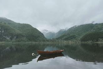 Wooden rowboat floating on a still lake surrounded by steep, forested mountains beneath an overcast sky