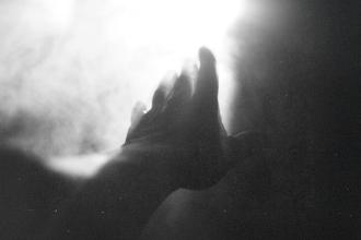 Hand reaching toward a bright light surrounded by soft, glowing mist in a black‑and‑white scene