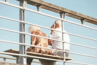 Two people sit on a bench outdoors behind a metal railing, with one holding a phone and clear blue sky in the background