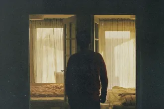 Silhouetted figure standing in a dimly lit doorway between two softly lit bedrooms with sheer curtains