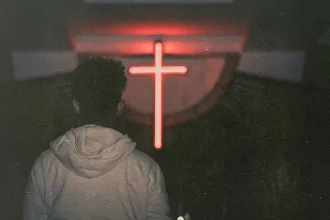 Silhouetted person standing in a dark room facing a glowing red cross on the wall