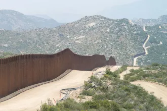 Long winding security wall running through a rugged, mountainous landscape with dirt roads following its path