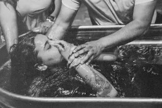 Two people assisting a woman being lowered into a water-filled baptism tub during a baptism ceremony