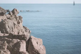 Rocky shoreline overlooking a calm blue sea with a distant sailboat on the horizon