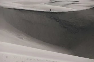 Person walking along the crest of a massive sand dune in a vast, windswept desert landscape