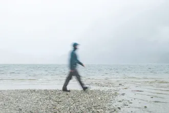 Person walking along a rocky lakeshore on a foggy day with mountains fading into the mist