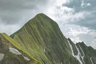 Steep green mountain peak with a small cross at the summit beneath a dramatic cloudy sky