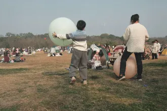 Children playing with large inflatable balls in an open grassy park filled with families, tents, and picnic groups on a clear day