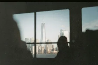 City skyline framed through a window, featuring a tall modern skyscraper and surrounding buildings with silhouetted observers in the foreground