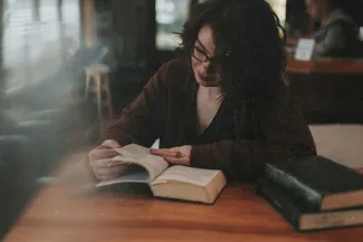 A person sits at a wooden table in a dimly lit room, reading an open book with another large book resting nearby