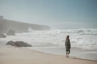 Person standing alone on a sandy beach, watching waves crash against a rocky shoreline under a clear blue sky