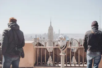 Two people standing at an observation deck overlooking the New York City skyline, with the Empire State Building centered in the view and a coin‑operated viewer in the foreground