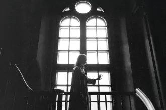 Silhouetted person standing indoors and touching a tall, bright window with arched panes, with light streaming in behind them