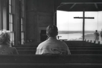 Two people sitting in church pews facing a large window with a cross silhouetted against a distant landscape, while others stand near the front