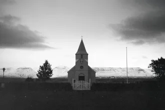 Small country church with a steeple standing before snow‑covered mountains at dusk in a black‑and‑white landscape