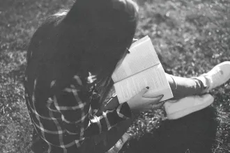 Person sitting on grass outdoors, holding an open book while reading in soft natural light