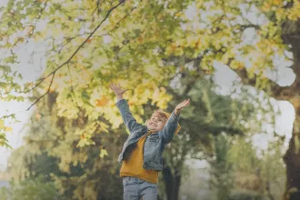 Child standing under a tree with autumn leaves, reaching upward in a sunlit park