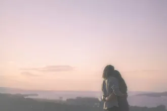Two people standing close together and embracing while looking out over a hazy landscape at sunset, with soft pastel colors in the sky