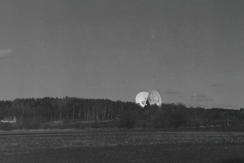 Large satellite dishes rising above a dense tree line in a wide, open field under a dark sky