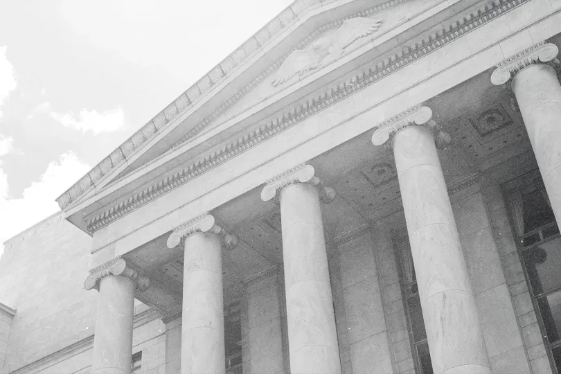 Low-angle black-and-white view of a large neoclassical building with tall columns and a triangular pediment against a cloudy sky