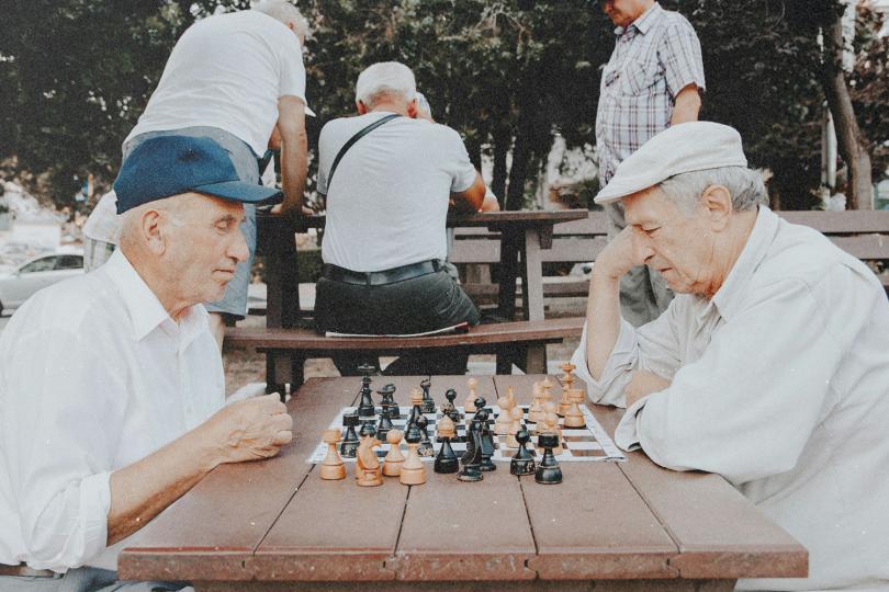 Two elderly males sit across from each other at a wooden picnic table outdoors playing a game of chess