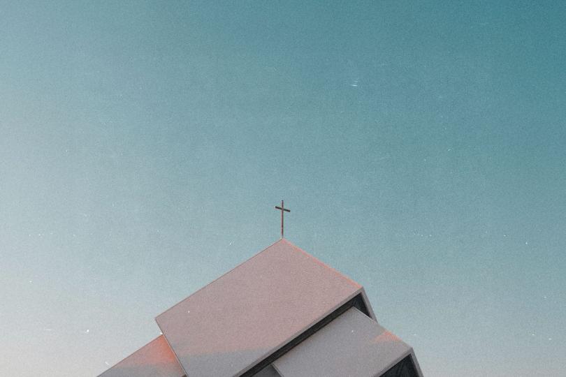 Low-angle view of a modern church roof with angular lines and a cross at the peak, set against a clear blue sky with subtle film grain texture