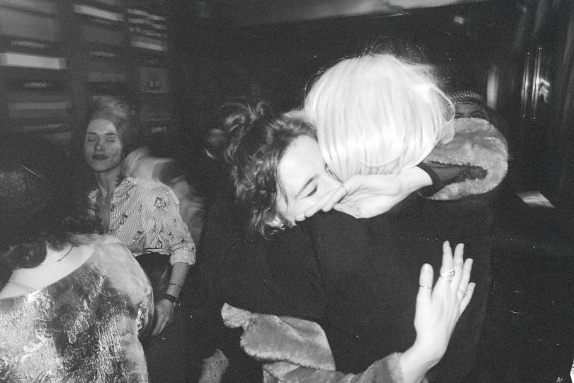 Two people embracing tightly in a crowded indoor space with others gathered and talking in the background