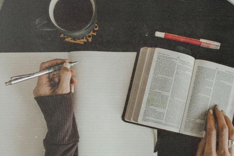 Open Bible and blank notebook on a table with a hand writing beside a cup of coffee