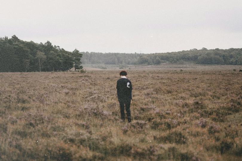 Person standing alone in a wide, grassy field bordered by distant trees under an overcast sky