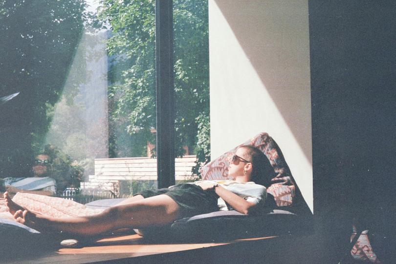 Person reclining on cushions beside a large window with sunlight streaming in and greenery outdoors